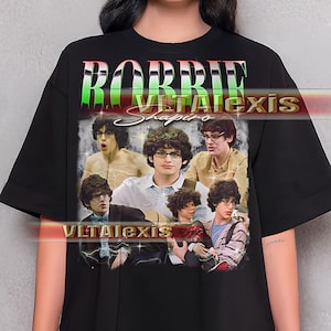 May include: Black t-shirt featuring a collage of images with the text "ROBBIE VITALEXIS Shapiro" in a green and red gradient font. The shirt has a vintage aesthetic, with multiple photos of a person wearing glasses.