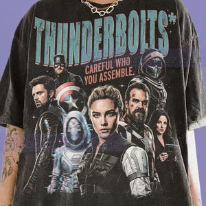 May include: Dark gray graphic t-shirt featuring the word "THUNDERBOLTS" in teal and a group of characters. The text "CAREFUL WHO YOU ASSEMBLE" is below the main title. The shirt is made of Comfort Colors Graphite material.
