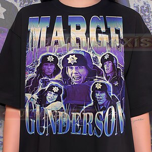 May include: Black t-shirt with a vintage-style graphic. The design features the text "MARGE GUNDERSON" in a distressed font, along with images of characters wearing hats with star badges, set against a purple and blue backdrop.