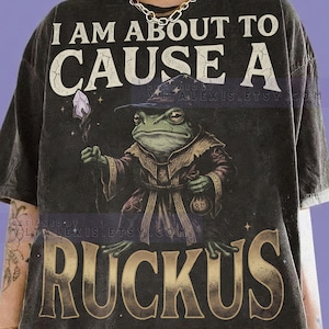 May include: Dark grey t-shirt with the text "I AM ABOUT TO CAUSE A RUCKUS." The shirt features a detailed illustration of a frog dressed as a wizard holding a staff and a bag. The shirt is made of Comfort Colors Graphite.