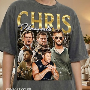 May include: Dark gray t-shirt featuring a graphic of Chris Hemsworth with the text "CHRIS Hemsworth" in gold and blue. The shirt also has the text "COMFORT COLOR PEPPER" at the bottom. The design includes multiple images of the actor.