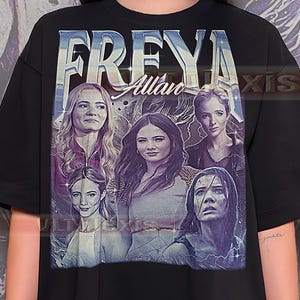Może przedstawiać: Czarny t-shirt z grafiką w stylu vintage. Projekt przedstawia napis "FREYA Allair" oraz portrety pięciu kobiet. Paleta kolorów obejmuje odcienie niebieskiego i fioletowego, ze efektem postarzenia.
