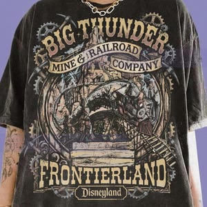 May include: A charcoal grey short-sleeve t-shirt featuring a graphic print of a steam train on tracks, gears, and pickaxes. The text reads BIG THUNDER MINE & RAILROAD COMPANY, FRONTIERLAND, Disneyland, and COMFORT COLORS GRAPHITE in various fonts.