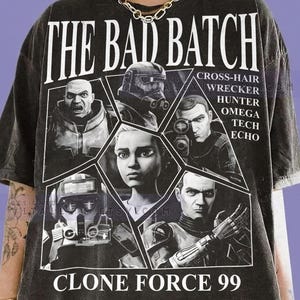 May include: Dark gray t-shirt featuring the text "THE BAD BATCH" and "CLONE FORCE 99". The shirt displays images of characters from the animated series, with names like Crosshair, Wrecker, and Hunter. The shirt is made of Comfort Colors Graphite material.