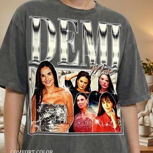 May include: A gray comfort color t-shirt featuring a graphic of Demi Moore with the word "DEMI" in large, silver, block letters. The word "Movie" is below the name. The text "COMFORT COLOR PEPPER" is printed at the bottom of the shirt.