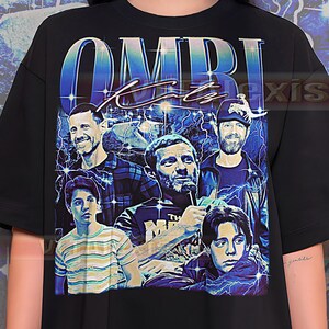 May include: Black t-shirt featuring a graphic of a group of people with the text "OMRI Katz" in a blue, retro-style design. The graphic includes images of the people and lightning bolts.