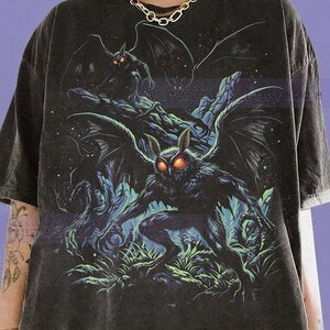 Könnte beinhalten: Graphitfarbenes T-Shirt mit einer detaillierten Illustration einer mottenartigen Kreatur mit leuchtend roten Augen und großen Flügeln. Das Design umfasst weitere ähnliche Figuren in einer dunklen, waldähnlichen Umgebung. Der Text "COMFORT COLORS" und "GRAPHITE" befindet sich unten.
