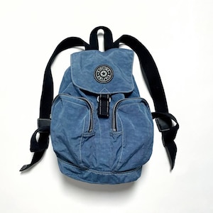 Könnte beinhalten: Blauer Nylon-Rucksack mit schwarzen Trägern und Reißverschlüssen. Der Rucksack hat einen Klappenverschluss mit einem runden Logo-Patch. Zwei Reißverschlusstaschen befinden sich auf der Vorderseite. Der Beutel befindet sich auf weißem Hintergrund.