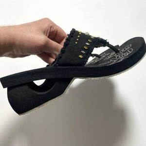 Könnte beinhalten: Schwarze Plateau-Flip-Flops mit dekorativem Riemen. Der Riemen besteht aus schwarzem Stoff mit ausgefransten Kanten und ist mit goldenen und silbernen Nieten verziert. Die Sohle hat einen weißen Rand und ein gedrucktes Design. Die Flip-Flops werden von einer Hand gehalten.