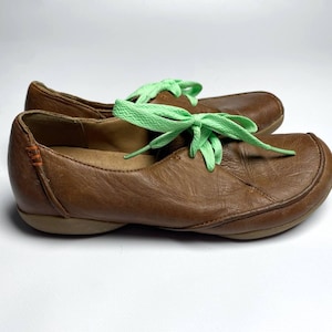 May include: A pair of brown leather shoes with light green laces. The shoes have a rounded toe and a low heel. The laces are tied in a bow. The shoes are on a white background.