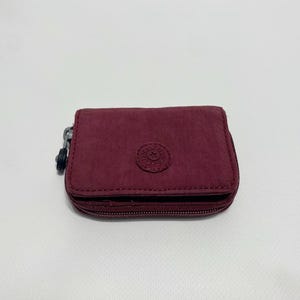 Peut inclure: Un portefeuille zippé bordeaux avec un petit logo circulaire au centre.