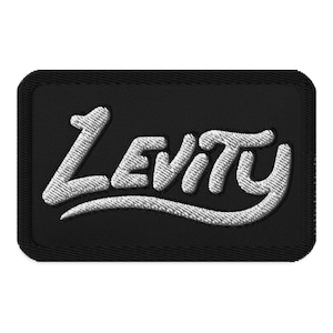 May include: Black embroidered patch with the word "Levity" in white, cursive lettering. The patch has a rectangular shape with rounded corners and a black border. The lettering is in a stylized, flowing font.