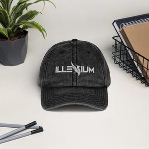 May include: A black vintage washed denim baseball cap with a white embroidered logo that says "ILLUSIUM" with a stylized bird design.