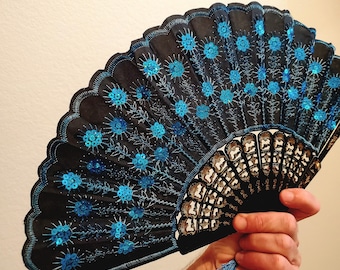 以下が含まれることがあります： 青いスパンコールと刺繍の花柄が施された黒い扇子。扇子はスカラップエッジと青いタッセルが付いています。扇子は開かれており、デザインの複雑な詳細が表示されています。