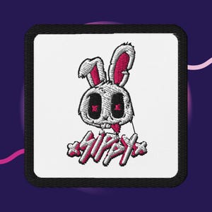 Puede incluir: Un parche bordado cuadrado con fondo blanco y borde negro. El diseño presenta una cabeza de conejo blanca con el interior de las orejas rosa y ojos en forma de X rosa. Debajo del conejo, el texto SIPPY está bordado en blanco con detalles rosas.
