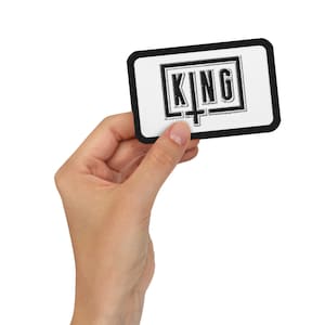 May include: Black and white embroidered patch with the word "KING" in a black square with a cross underneath. The patch is held in a hand against a white background.