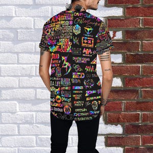 Può includere: T-shirt nero con un collage di loghi e testi colorati, tra cui nomi di artisti come David Guetta e Calvin Harris. Il design è vivace e accattivante, con una varietà di font ed elementi grafici. La persona indossa pantaloni neri.