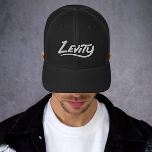 May include: Black trucker cap with a curved peak and mesh back. The word "Levity" is embroidered in white script across the front. The cap is worn by a person.