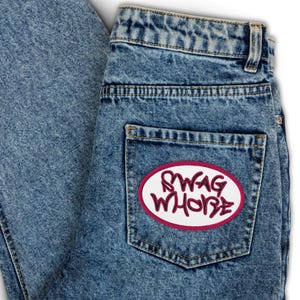 May include: Acid-wash blue denim jeans featuring a high-waisted design with a back pocket. An oval-shaped patch is centered on the pocket, featuring a white background, a dark pink border, and the black text SWAG WHORE in a stylized font.