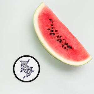 Op de afbeelding: Een witte ronde patch met een zwarte rand en een zilveren geborduurd ontwerp van de letters "ZD" met prikkeldraad eromheen. Op de achtergrond is een plak watermeloen te zien.