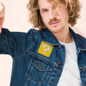 May include: A man wearing a blue denim jacket with a yellow question mark block patch on the left side of the chest.