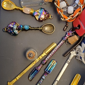 Op de afbeelding: Een assortiment decoratieve lepels en schepjes in verschillende ontwerpen en kleuren. Sommige lepels hebben sierlijke handvatten met edelstenen en draadwikkelingen. Andere schepjes hebben iriserende afwerkingen en schedeldetails. Een klein potje met een kurken deksel is ook zichtbaar.