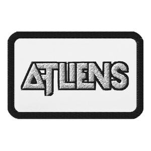 May include: Embroidered patch with the word "ATHENS" in a stylized, 3D effect font. The text is in silver against a white background, framed by a black border. The patch is rectangular with rounded corners.