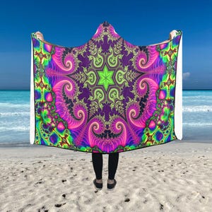 Pode incluir: Uma manta com capuz com um design psicodélico vibrante. A manta apresenta um padrão central de estrela em verde néon, rodeado por formas fractais rodopiantes em tons de roxo, rosa e azul. O fundo é uma cena de praia com céu azul e oceano.