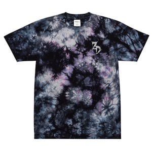 May include: A tie-dye t-shirt in shades of black, purple, and white. The shirt features a small, stylized silver emblem on the chest. The fabric has a textured, swirling pattern.