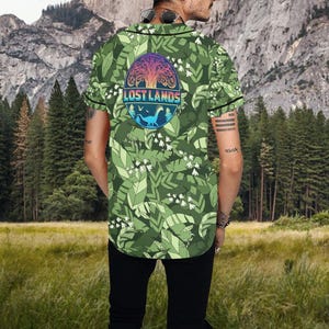 May include: Green baseball jersey with a leafy pattern and the text "LOST LANDS" in a circular design. The design includes a tree, a dinosaur, and a mountain range. The jersey has black trim and short sleeves.