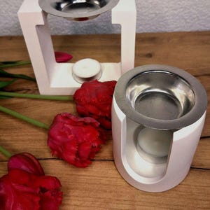 Könnte beinhalten: Zwei weiße Diffusoren für ätherische Öle mit silbernen Schalen, einer in einem quadratischen Rahmen und der andere in runder Form. Rote Tulpen sind auf einer Holzoberfläche angeordnet. In jedem Diffusor befindet sich eine brennende Kerze.
