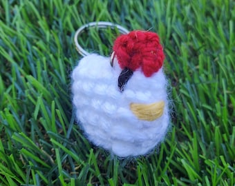 May include: A handmade white chicken keychain with a red comb and yellow beak. The keychain is made of a soft, textured material and has a silver metal ring. The chicken is set against a background of green grass.