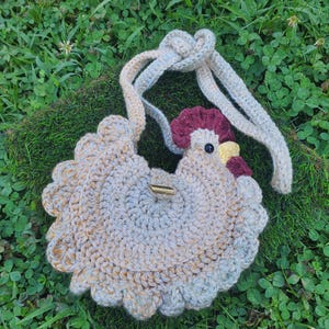 May include: A handmade crochet purse shaped like a hen, with a button closure. The purse features a light beige body with a scalloped edge, a red comb, and a yellow beak. The strap is a light beige color. The purse is lying on a bed of green grass.