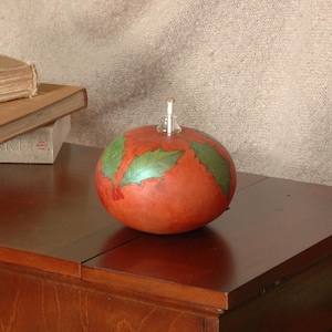 Pode incluir: Uma cabaça vermelha com folhas verdes de azevinho pintadas sobre ela, usada como uma lâmpada de óleo decorativa. A lâmpada está sentada em uma mesa de madeira com livros empilhados sobre ela.
