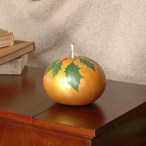 Pode incluir: Um porta-velas de abóbora dourado com um design de folhas de azevinho verdes. O porta-velas está sobre uma mesa de madeira.