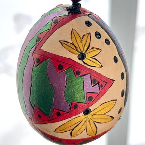 Peut inclure: Pendentif œuf décoratif peint à la main avec un motif géométrique. L'œuf présente une base beige avec des accents colorés verts, violets, rouges et dorés. Deux motifs de fleurs dorées sont peints sur la surface. Suspendu à un cordon marron foncé.