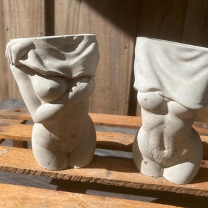 May include: Two concrete sculptures of a torso with a fabric draped over the top. The sculptures are a light gray color and have a rough texture.