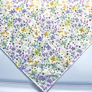 Puede incluir: Una tela blanca con un estampado floral repetido. El diseño presenta flores moradas, amarillas y azules con hojas verdes y pequeñas bayas moradas oscuras. Las flores están dispersas por la tela.