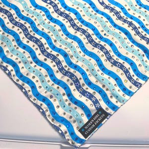 Puede incluir: Una tela cuadrada estampada con rayas onduladas en tonos azules y blancos, adornada con pedrería plateada y azul y detalles en forma de estrella. La tela tiene una etiqueta que dice "MAMA'S GIRL CUSTOM JACKETS".