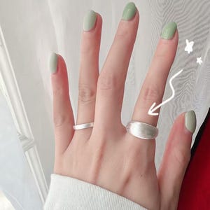May include: A hand wearing two silver rings. The ring on the index finger is a simple band, while the ring on the middle finger is a wider band with a rounded top.