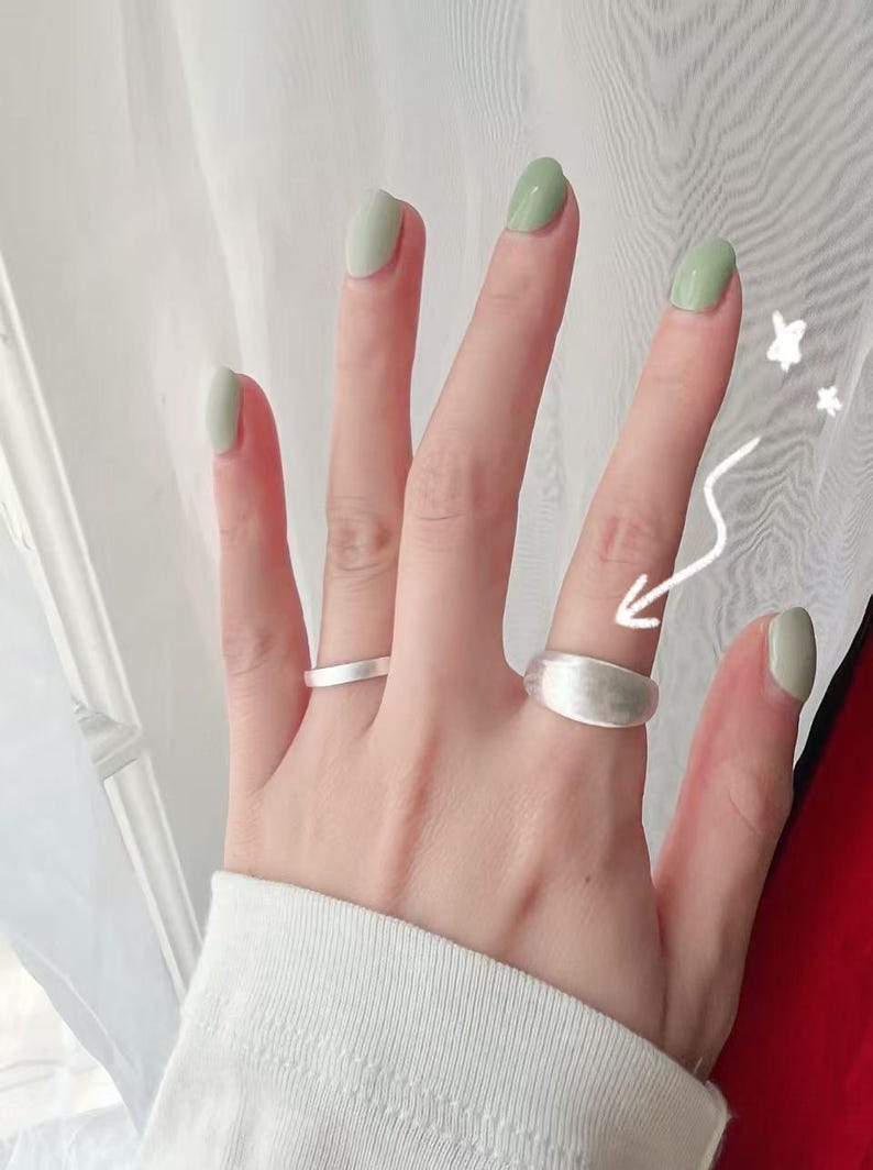 May include: A hand wearing two silver rings. The ring on the index finger is a simple band, while the ring on the middle finger is a wider band with a rounded top.