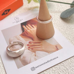 May include: Two silver rings on a wooden ring display stand. The rings are both simple bands with a brushed finish. The stand is a cone shape with a light brown color.