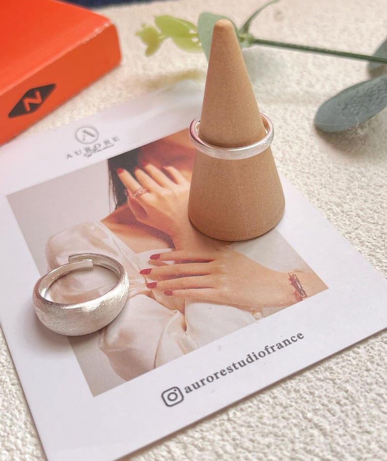 May include: Two silver rings on a wooden ring display stand. The rings are both simple bands with a brushed finish. The stand is a cone shape with a light brown color.