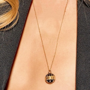 Puede incluir: Un collar dorado con un colgante esférico. El colgante es negro con detalles dorados y pequeñas piedras transparentes. El collar cuelga sobre el pecho de una persona, con una chaqueta oscura visible a los lados.