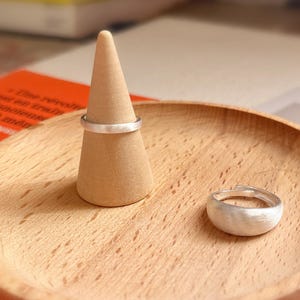May include: Two silver rings on a wooden ring holder. The ring on the left is a thin band, and the ring on the right is a wider band with a textured surface.