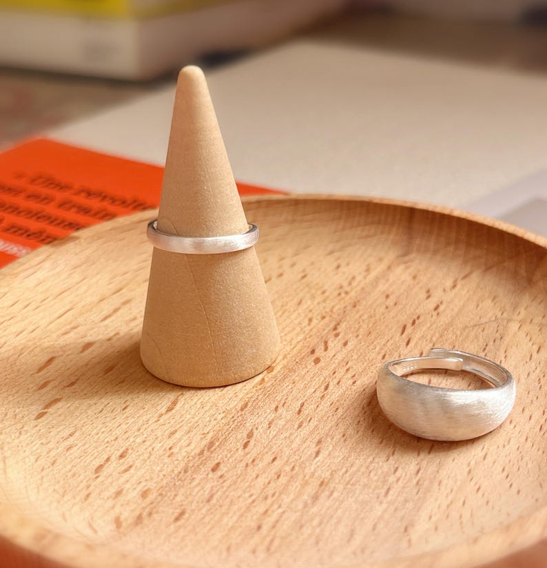 May include: Two silver rings on a wooden ring holder. The ring on the left is a thin band, and the ring on the right is a wider band with a textured surface.