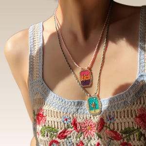 Puede incluir: Dos collares con colgantes rectangulares. Un colgante tiene el texto "Love is the Answer" con un diseño de puesta de sol. El otro colgante tiene un diseño de palmera. Ambos colgantes están en cordones de cuentas coloridas.