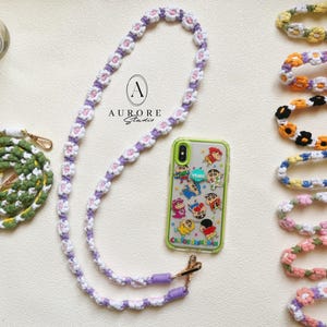 Pode incluir: Uma coleção de alças de telefone feitas à mão com flores de crochê em várias cores, incluindo uma alça longa de margaridas lavanda e brancas presa a uma capa transparente com personagens e o texto AURORE Studio.