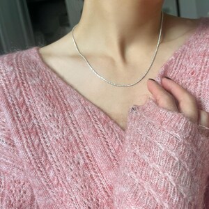 Puede incluir: Un delicado collar de plata con una cadena texturizada se lleva sobre un suéter rosa. El collar es simple y elegante, reflejando la luz. El suéter tiene una textura suave y difusa y un cuello en V.