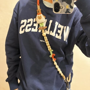 May include: A macrame phone strap with a beige and orange woven design. The strap features a small bear-shaped charm wearing a striped shirt and green pants. The strap is attached to a navy blue sweatshirt with the word "WETLESS" printed on it.